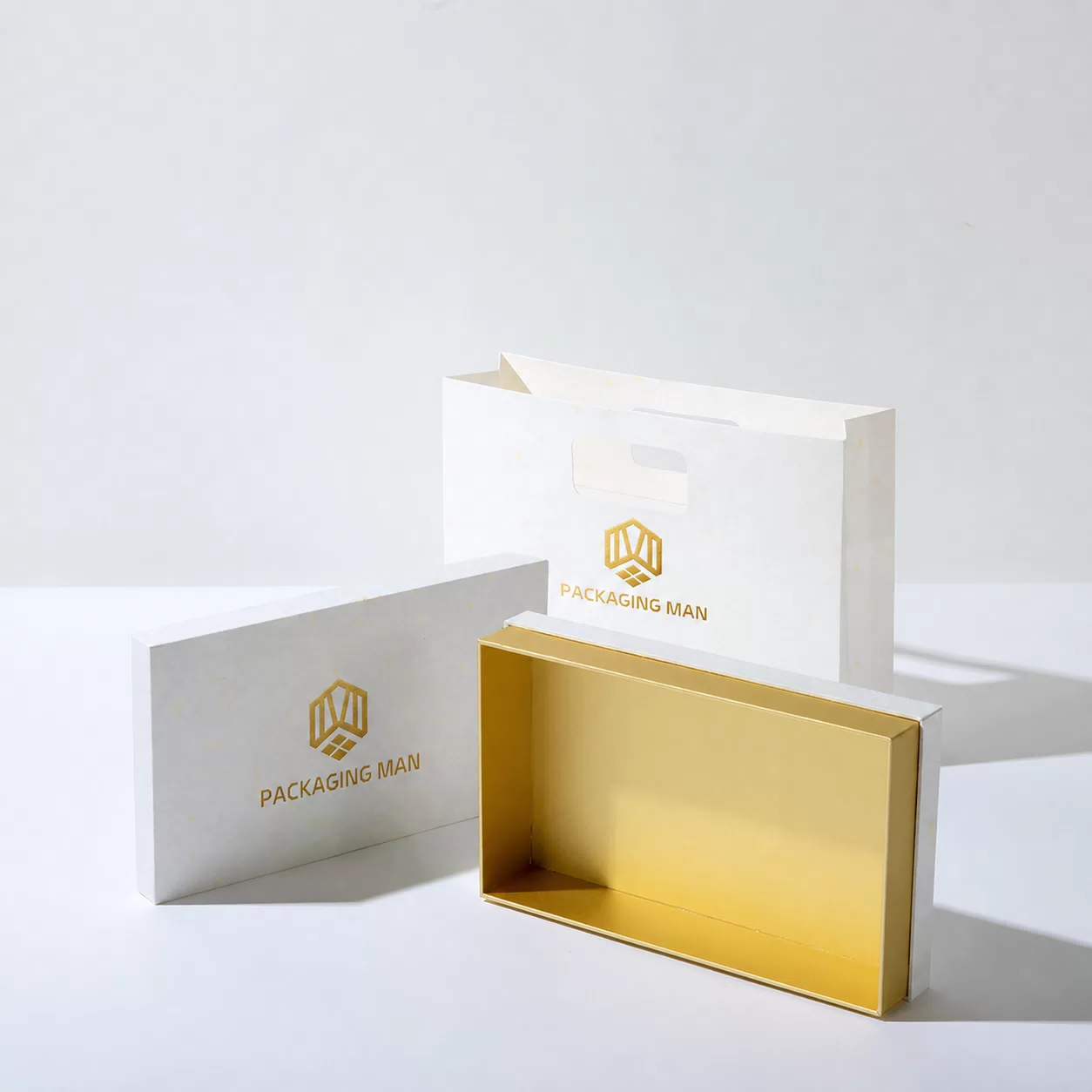 The white box is shown open with its lid removed, revealing a shiny gold interior base, while the white bag stands in the background.